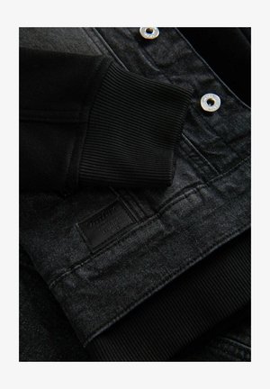 Black denim jacket detail with textured fabric, ribbed cuffs, silver snaps, and a black label featuring "Ombre" embossed at the hem.