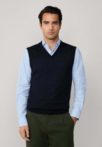 Muž v čiernej bezrúkavovej sveteri s V-výstrihom, pod ktorým má svetlomodrú košeľu s gombíkmi a tmavoolivové nohavice, stojí so zapichnutou jednou rukou v vrecku.