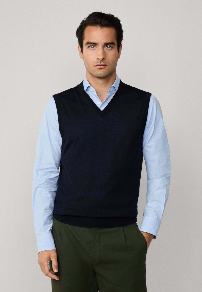 Muž v čiernej bezrúkavovej sveteri s V-výstrihom, pod ktorým má svetlomodrú košeľu s gombíkmi a tmavoolivové nohavice, stojí so zapichnutou jednou rukou v vrecku.