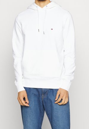 Mann trägt einen schlichten weißen Hoodie mit Bauchtasche und kleinem Logo auf der Brust, kombiniert mit blauen Jeans, steht vor einem einfarbigen Hintergrund.