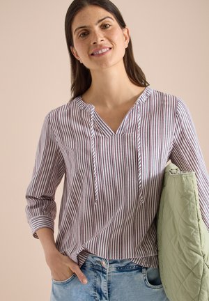 Mujer sonriendo, vistiendo una camiseta de manga larga a rayas y pantalones vaqueros azul claro, sosteniendo una chaqueta verde acolchada doblada bajo el brazo.