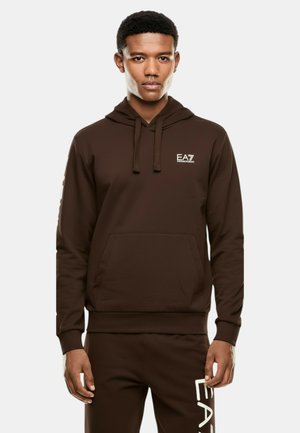 Giovane in felpa marrone con cappuccio e pantaloni abbinati, con logo bianco EA7 sul petto e sulla gamba sinistra, in piedi su uno sfondo neutro.
