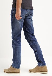 Hombre caminando con jeans azul desgastados, camiseta negra y zapatos de ante color beige frente a un fondo blanco liso.