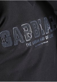 Sudadera negra con tejido texturizado, que presenta grandes letras en relieve "GABBIANI" y "EL ESTILO DE VIDA URBANO" en colores contrastantes.