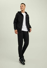 Schwarze Zip-Hoodie mit Kordelzügen, kombiniert mit schwarzen Jogginghosen. Darunter ein weißes T-Shirt. Getragen mit schwarzen Sportschuhen mit gemustertem Detail.