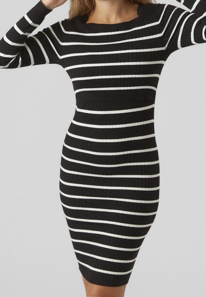 Femme portant une robe noire moulante à rayures blanches horizontales et manches longues, se tenant devant un arrière-plan gris clair uni.