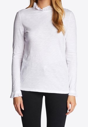 Haut blanc à manches longues avec un col à volants et une texture subtile. Présente des poignets à volants, ici associé à un pantalon noir.