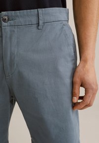 Svetlo sivi bombažni hlače s ravnim krojem, z zapiranjem na gumb, zankami za pas in gladko teksturo. Roka je postavljena na strani.