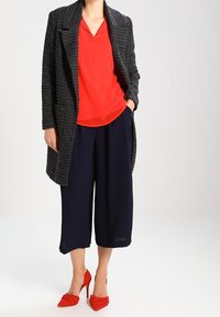Manteau long texturé noir, top rouge à col en V, pantalons culottes bleu marine et chaussures à talons rouges avec un détail en nœud.