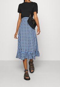 Person, die ein schwarzes kurzärmliges Oberteil, einen blau-schwarz karierten Midijupf mit Rüsche am Saum, schwarze Sandalen mit Schnallen und eine schwarze Umhängetasche trägt.