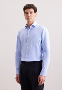 Lichtblauwe overhemd met lange mouwen van gladde stof, voorzien van een knoopsluiting aan de voorkant en een borstzak. G gedragen met donkere broek.