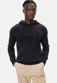 Felpa con cappuccio blu navy in tessuto liscio, con un logo "GUES" in rilievo tono su tono all'interno di un triangolo ben visibile. Polsini e orlo aderenti.