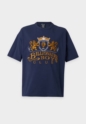 Młody mężczyzna w niebieskim t-shircie Billionaire Boys Club na koszuli w kratę, jasnoniebieskich dżinsach, czarnych butach, czapce moro i trzymający wzorzystą torbę.