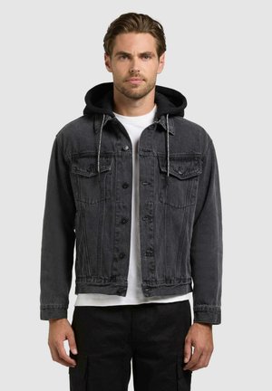 Mand iført sort denimjakke med sort hættetrøje under og hvid skjorte, stående foran ensfarvet baggrund.