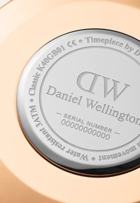 Fondo circular de reloj de plata con texto grabado, incluyendo la marca "Daniel Wellington", número de serie y resistencia al agua hasta 30 metros, sobre un fondo rosa dorado.