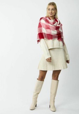Châle à carreaux rouges et roses sur un pull en tricot crème et une jupe plissée ; mannequin portant des bottes beiges à talons jusqu'aux genoux.
