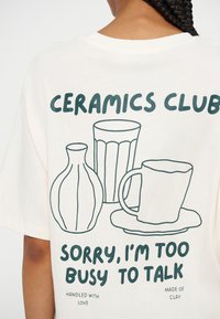 Човек, който носи бяла риза с текст "Ceramics Club" и прости илюстрации на ваза, чаша и чиния на гърба.
