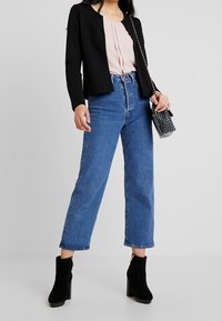 Czarna strukturalna marynarka na różowej bluzce z pliskami, w połączeniu z niebieskimi dżinsami o wysokim stanie i czarnymi botkami na obcasie. W zestawie mała czarna torebka.