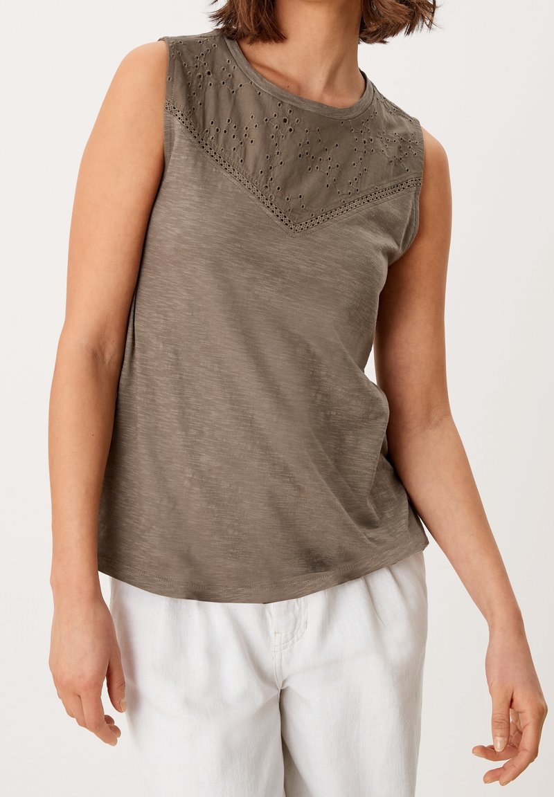 Haut sans manches vert olive avec empiècement en dentelle à œillets, associé à un pantalon blanc ample sur une personne debout devant un fond uni.