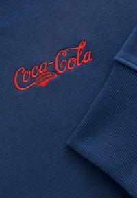 Tissu bleu marine avec le logo "Coca-Cola" brodé en rouge et des détails d'accent. Présente des poignets côtelés et une texture lisse, mettant l'accent sur la marque.