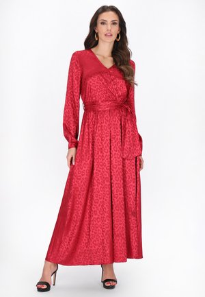 Žena v červených šatech s dlouhým rukávem a zavinovacím střihem s jemným leopardím vzorem a černých sandálech na vysokém podpatku na bílém pozadí.