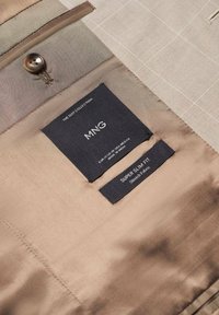Beige colbertvoering met een enkele knoop, een zak en twee labels met de tekst "MNG" en "SUPER SLIM FIT", met een gladde textuur.