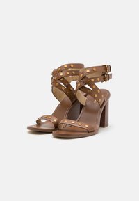 Brune høye hælsandaler med naglede stropper, ankelspenner og en strukturert blokkheel. Åpen tå-design med glatt skinnfinish.