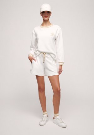 Sudadera blanca con detalles y logotipo en dorado, combinada con pantalones cortos blancos con cordones. Completado con zapatillas blancas y una gorra a juego.