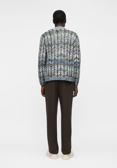 Άνδρας που φοράει πολύχρωμο πουλόβερ με ζιγκ-ζαγκ μοτίβο, σκούρο καφέ παντελόνι και λευκά αθλητικά παπούτσια, στέκεται με την πλάτη προς τα εμπρός σε απλό φόντο.