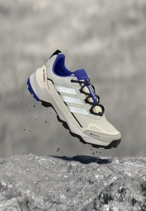Zapatilla de senderismo Adidas Terrex Gore-Tex en azul y gris claro suspendida en el aire sobre terreno rocoso con partículas de tierra cayendo.