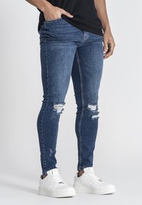 Jeans de denim azul ajustados con desgastes y roturas, que presentan una cintura media y un diseño ceñido, combinados con zapatillas blancas.