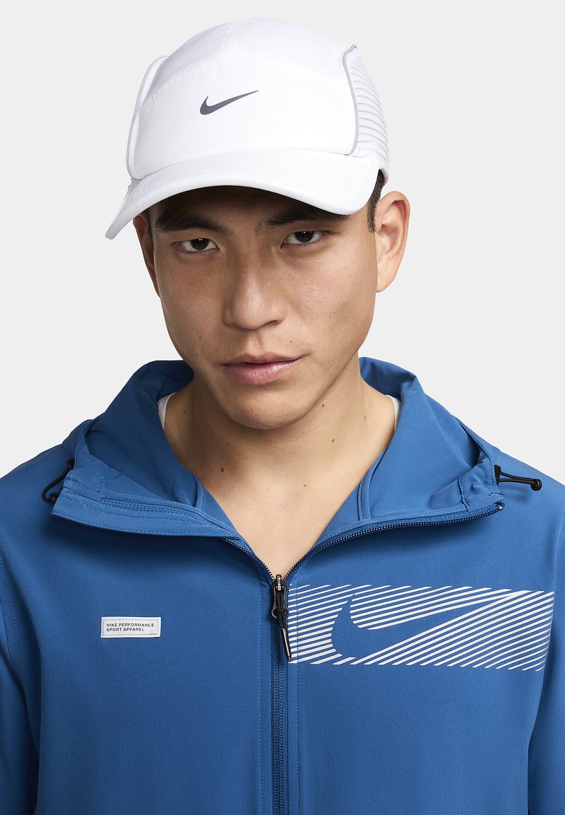 Hvid Nike-cap med buet skygge og grå logo, båret af en person i en blå zip-up jakke med et stribet grafik og en logotag.