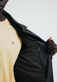 Giacca di pelle nera, con cerniera e una finitura liscia, indossata sopra una maglietta gialla con un piccolo ricamo di un albero verde.