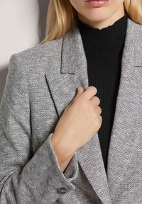 Grauer Fischgrätblazer mit Revers und geknöpften Manschetten. Über einem schwarzen, gerippten Rollkragenoberteil getragen. Weiche Textur und taillierte Passform.