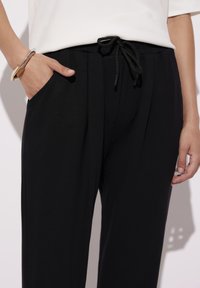 Czarne spodnie wykonane z miękkiego materiału, z zakładkami, elastycznym pasem z regulowanym troczkiem i kieszeniami po bokach.