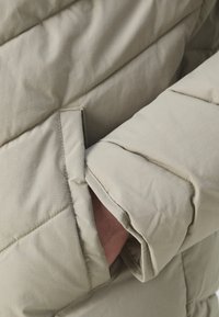 Manche de veste matelassée gris clair avec des coutures diagonales, une poche cachée et une doublure intérieure douce. Le matériau semble lisse et texturé.