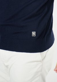 Pull en tricot marine avec un ourlet côtelé et un petit logo en bas à gauche. Texture douce, design ajusté, associé à un pantalon blanc.