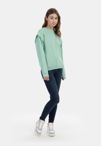 Felpa verde menta con cuciture sulle spalle scese, abbinata a jeans skinny blu scuro e sneaker alte multicolori.
