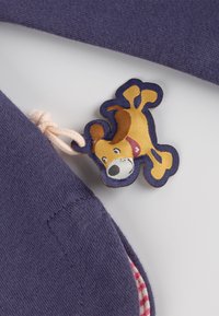 Purple fabric with a textured surface, featuring a tag of a cartoon dog in yellow and brown. The tag has a light pink drawstring.