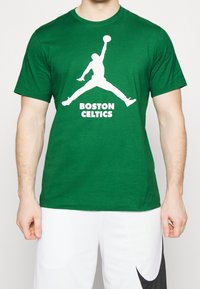 Mies, joka pukeutuu vihreään t-paitaan, jossa on valkoinen koripalloilijan siluetti ja teksti "Boston Celtics", parina valkoiset shortsit, joissa on musta swoosh.