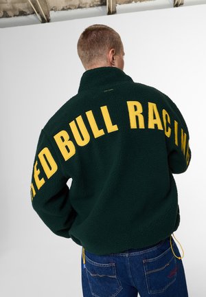Person trägt eine dunkelgrüne Fleecejacke mit großem gelben "Red Bull Racing"-Text auf dem Rücken und blaue Jeans, steht vor einer weißen Wand.
