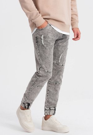 Gray distressed jeans with a tapered fit, featuring ripped detailing and an adjustable cuff. Paired with white sneakers and a beige sweatshirt.