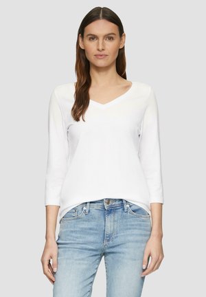 Top a maniche lunghe bianco con scollatura a V realizzato in tessuto morbido, abbinato a jeans azzurri chiari. Il top ha una vestibilità comoda e presenta dettagli cuciti.