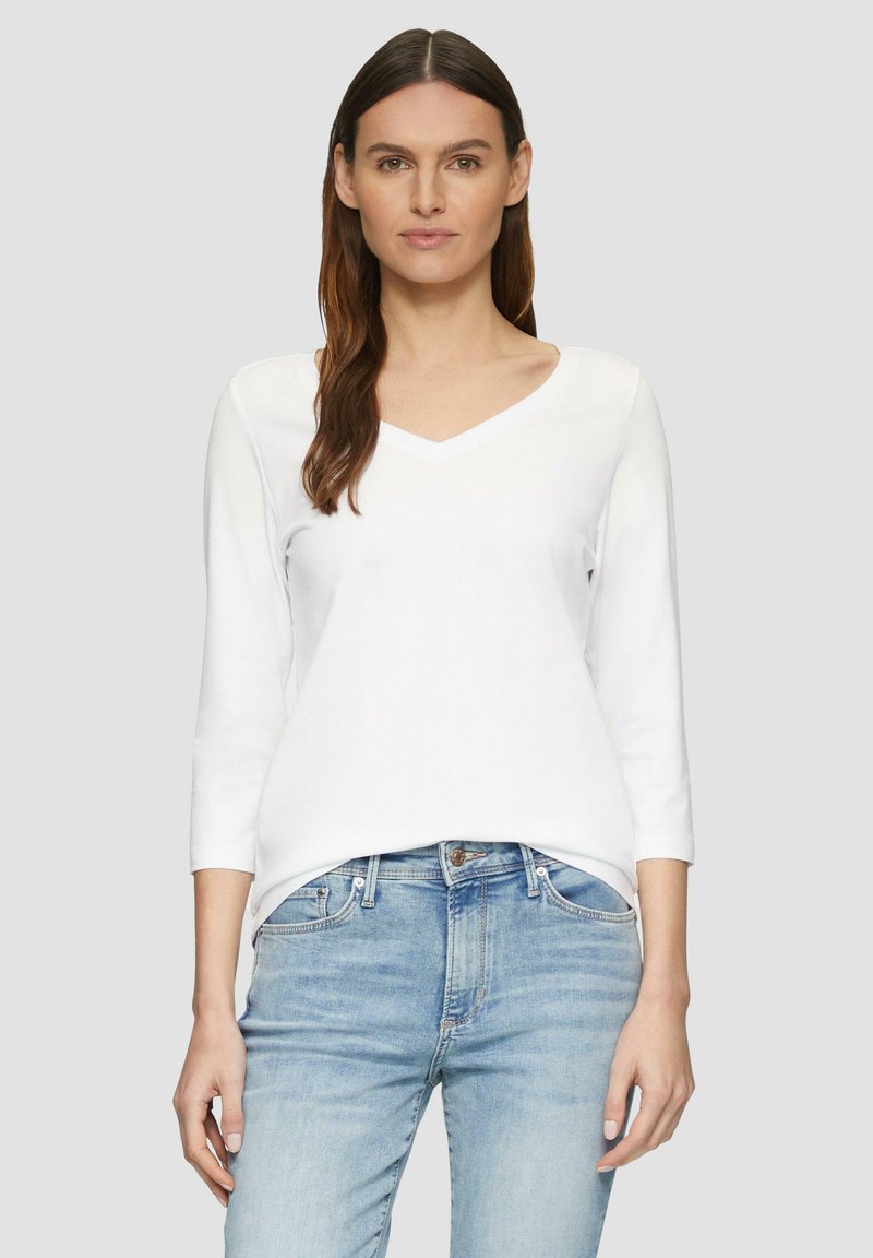 Witte V-hals top met lange mouwen van zacht fabric, gecombineerd met lichtblauwe jeans. De top heeft een relaxed fit en beschikt over gestikte accenten.