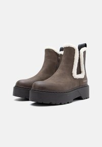 Botas de tobillo marrones con suelas negras gruesas, cremalleras laterales y un borde de pelo sintético blanco alrededor del tobillo, además de tiradores en la parte trasera.
