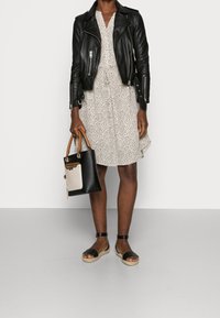 Schwarze Leder-Bikerjacke über einem weißen Kleid mit schwarzen Sprenkeln. Hellbraune und schwarze Handtasche mit einem weißen Akzent. Schwarze Sandalen mit Knöchelriemen.