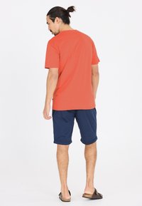 Röd kortärmad skjorta, marinblå shorts och sandaler. Skjortan har en rund halsringning och en avslappnad passform; shortsen är knälånga med uppvikta fållar.