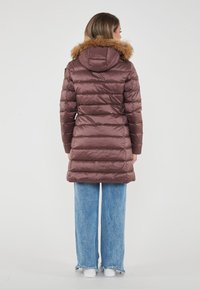 Donna che indossa un lungo cappotto imbottito marrone con cappuccio foderato di pelliccia, pantaloni jeans larghi blu e sneakers bianche, in piedi di spalle su uno sfondo bianco.