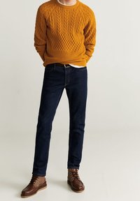Personne portant un pull en maille torsadée jaune moutarde, un jean bleu foncé et des bottes en cuir marron à lacets, debout les mains derrière le dos.
