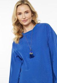 Kék bordázott pulóver laza szabással és hosszú ujjal, kék bojt nyaklánccal és arany karikafülbevalóval kiegészítve.